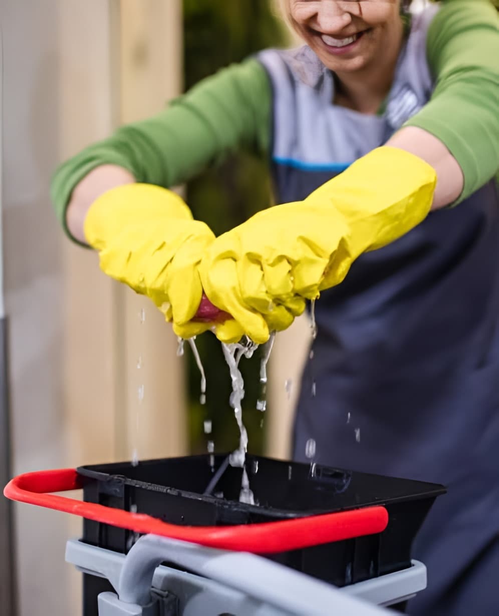 Handen met gele schoonmaakhandschoenen wringen een doek uit boven een emmer